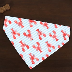 May include: A triangular dog bandana with a repeating pattern of red lobsters on a light blue and white striped background. The bandana is made of fabric and is designed to be worn around a dog's neck. Two dog treats are visible.