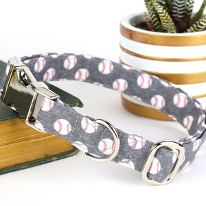 May include: A gray dog collar with a white and red baseball pattern. The collar has a silver buckle and a silver D-ring.