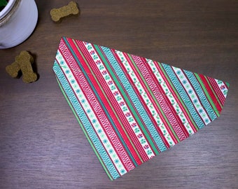Puede incluir: Un bandana para mascotas con estampado rojo, verde y blanco. El bandana tiene un tema navideño con rayas y copos de nieve.