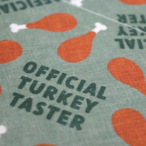 May include: Green fabric with the text "OFFICIAL TURKEY TASTER" in dark green letters. The fabric is decorated with a repeating pattern of orange turkey legs.