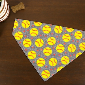 May include: A gray dog bandana with a pattern of yellow softballs and small red hearts. The softballs have red stitching details. The bandana is made of fabric and is designed to be worn around a dog's neck.