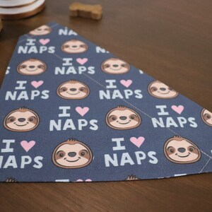 Puede incluir: Un pa&ntilde;uelo para perros azul marino con un patr&oacute;n repetido de perezosos sonrientes, corazones rosas y el texto "I ❤️ NAPS" en blanco. El pa&ntilde;uelo es triangular y de tela.