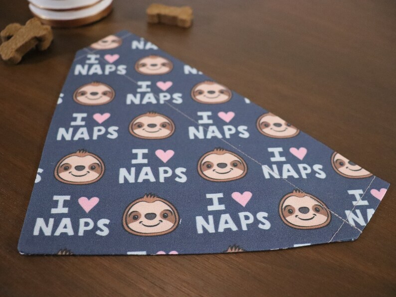 Puede incluir: Un pa&ntilde;uelo para perros azul marino con un patr&oacute;n repetido de perezosos sonrientes, corazones rosas y el texto "I ❤️ NAPS" en blanco. El pa&ntilde;uelo es triangular y de tela.