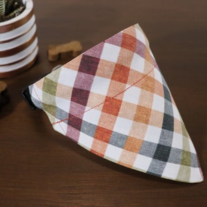 May include: A white, orange, green, and burgundy plaid dog bandana with a red stitched edge.