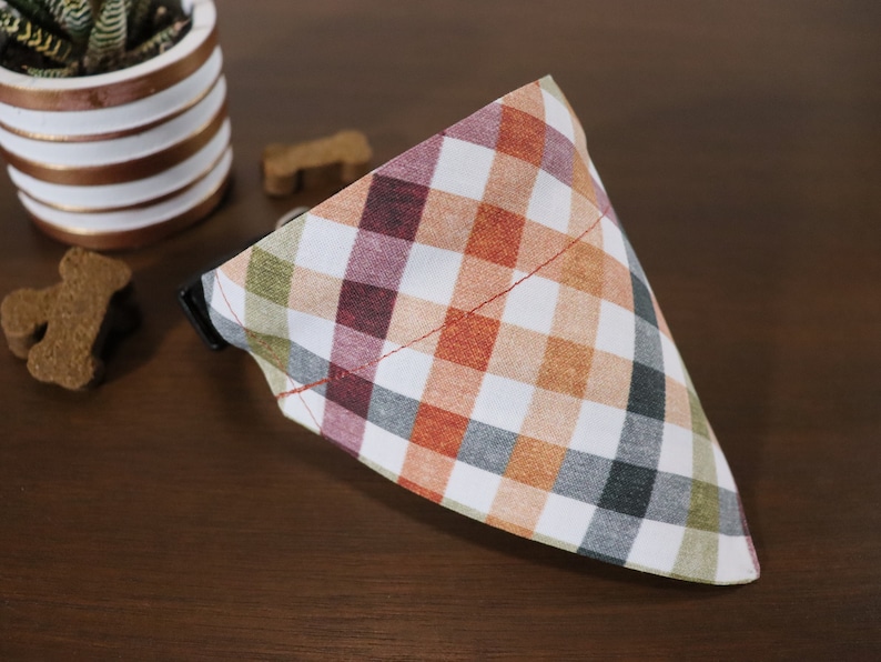 May include: A white, orange, green, and burgundy plaid dog bandana with a red stitched edge.