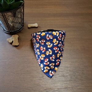 May include: A blue bandana with a pattern of orange, yellow, and brown foxes. The bandana is triangular and has a black adjustable strap.