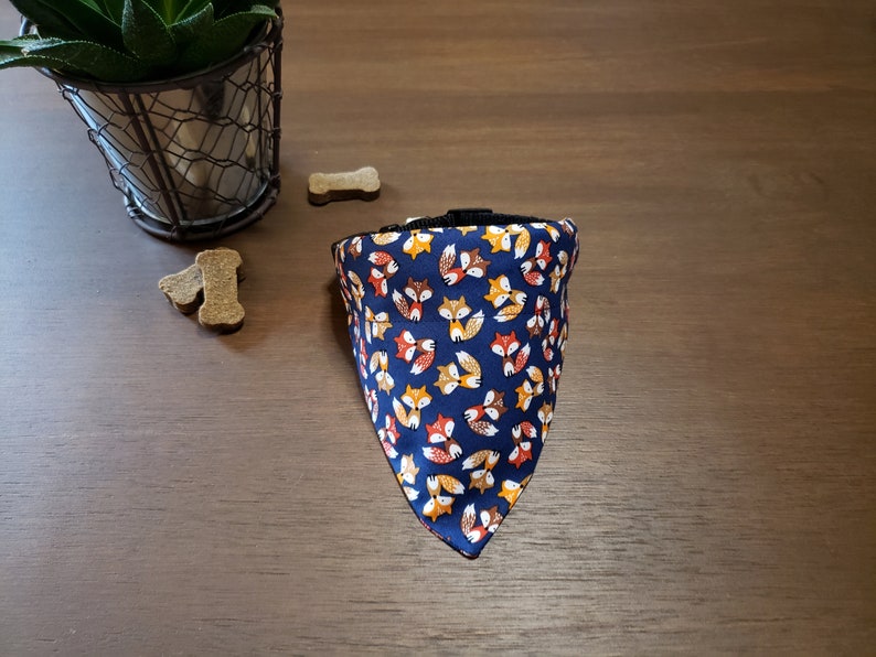May include: A blue bandana with a pattern of orange, yellow, and brown foxes. The bandana is triangular and has a black adjustable strap.