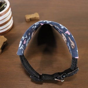 Puede incluir: Un collar para perros azul marino con una tela estampada con texto blanco, corazones rosas e ilustraciones de perros. El collar tiene una correa de nailon negro y una hebilla de pl&aacute;stico. Una peque&ntilde;a planta suculenta y golosinas para perros est&aacute;n en el fondo.