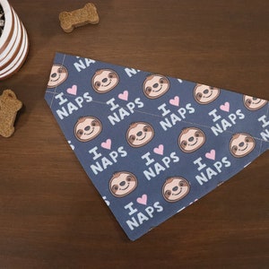 Puede incluir: Bandana para perros azul marino con un patr&oacute;n repetido de perezosos sonrientes, corazones rosas y el texto "I love naps". La bandana est&aacute; doblada sobre una superficie de madera, con golosinas para perros y una planta en maceta en el fondo.