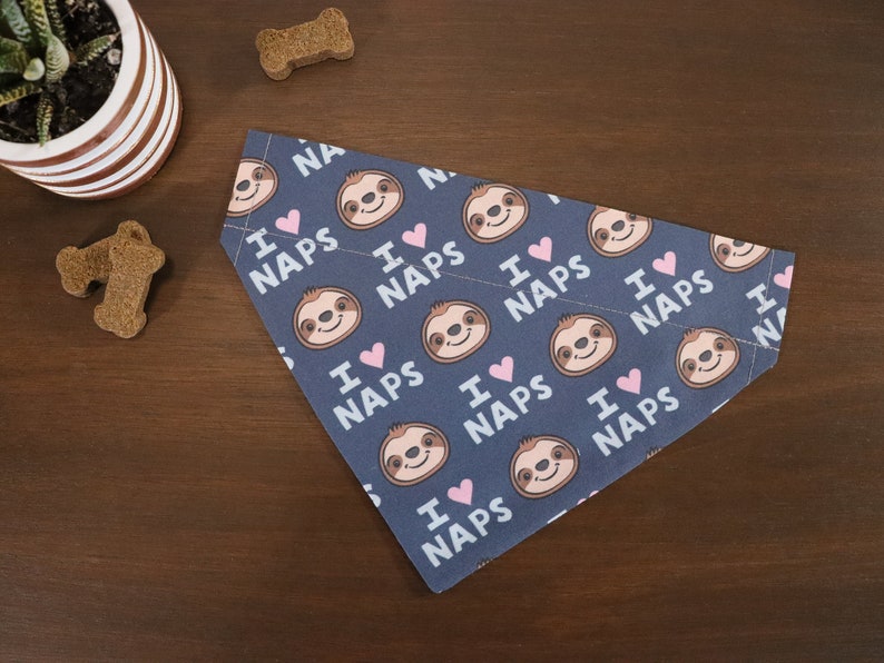 Puede incluir: Bandana para perros azul marino con un patr&oacute;n repetido de perezosos sonrientes, corazones rosas y el texto "I love naps". La bandana est&aacute; doblada sobre una superficie de madera, con golosinas para perros y una planta en maceta en el fondo.
