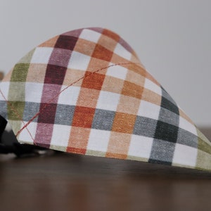 May include: A plaid dog bandana with a white background and orange, green, gray, and burgundy plaid pattern. The bandana has a black adjustable collar strap.
