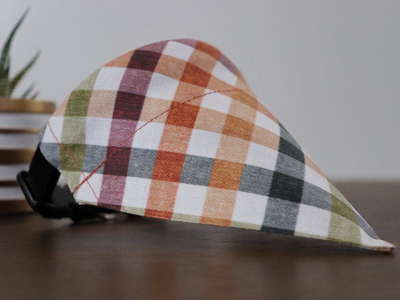 May include: A plaid dog bandana with a white background and orange, green, gray, and burgundy plaid pattern. The bandana has a black adjustable collar strap.