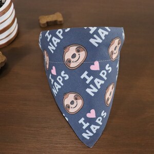 Puede incluir: Bandana azul para perros con un patr&oacute;n repetido de perezosos sonrientes, corazones rosas y las palabras "I ❤️ NAPS" en blanco. La bandana est&aacute; doblada sobre una superficie de madera, con golosinas para perros y una planta en maceta en el fondo.