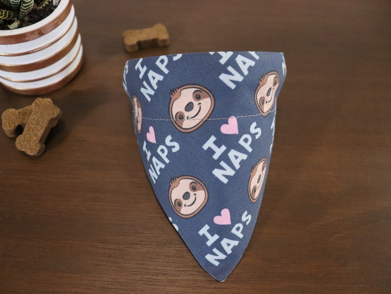 Puede incluir: Bandana azul para perros con un patr&oacute;n repetido de perezosos sonrientes, corazones rosas y las palabras "I ❤️ NAPS" en blanco. La bandana est&aacute; doblada sobre una superficie de madera, con golosinas para perros y una planta en maceta en el fondo.