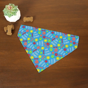 May include: A blue dog bandana with a repeating pattern of paw prints, bones, and suns. The text "HERE COMES TROUBLE!" is scattered across the fabric in white and yellow. The bandana is triangular in shape.