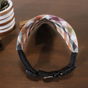 May include: A black dog collar with a plaid fabric pattern in shades of brown, green, and white. The collar has a black buckle and is adjustable.