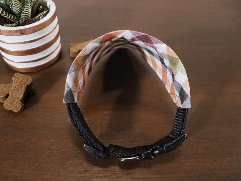 May include: A black dog collar with a plaid fabric pattern in shades of brown, green, and white. The collar has a black buckle and is adjustable.