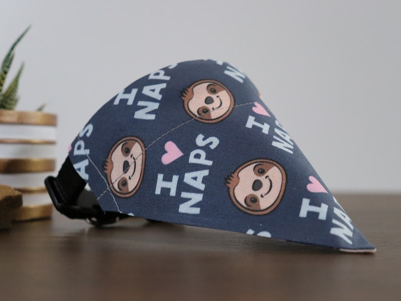 Puede incluir: Bandana azul marino para perros con un patr&oacute;n repetido de caras de perezosos sonrientes, corazones rosas y las palabras "I NAPS" en blanco. La bandana tiene un collar negro ajustable.