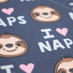 Puede incluir: Primer plano de una tela azul marino con un patr&oacute;n repetido de caras de perezosos sonrientes, corazones rosas y las palabras "I LOVE NAPS" en blanco. Las caras de los perezosos tienen pelaje marr&oacute;n y beige.