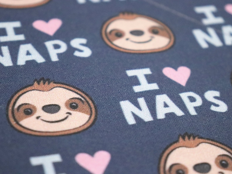 Puede incluir: Primer plano de una tela azul marino con un patr&oacute;n repetido de caras de perezosos sonrientes, corazones rosas y las palabras "I LOVE NAPS" en blanco. Las caras de los perezosos tienen pelaje marr&oacute;n y beige.