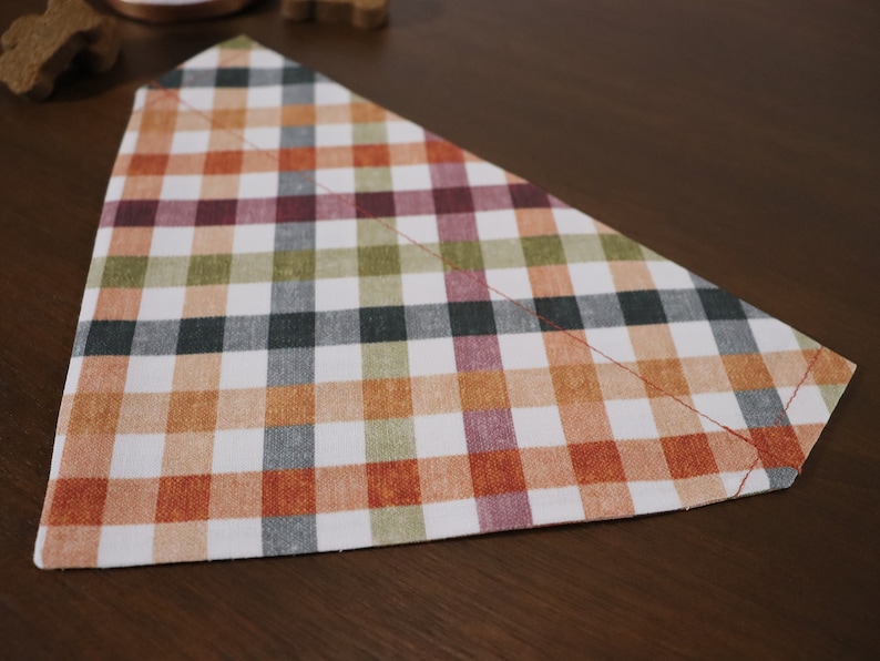 May include: A triangular dog bandana with a plaid pattern in shades of orange, green, black, and pink.