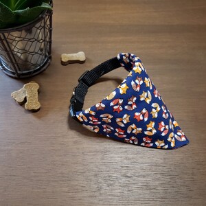 May include: A blue dog bandana with a pattern of orange, yellow, and brown foxes. The bandana is attached to a black collar.