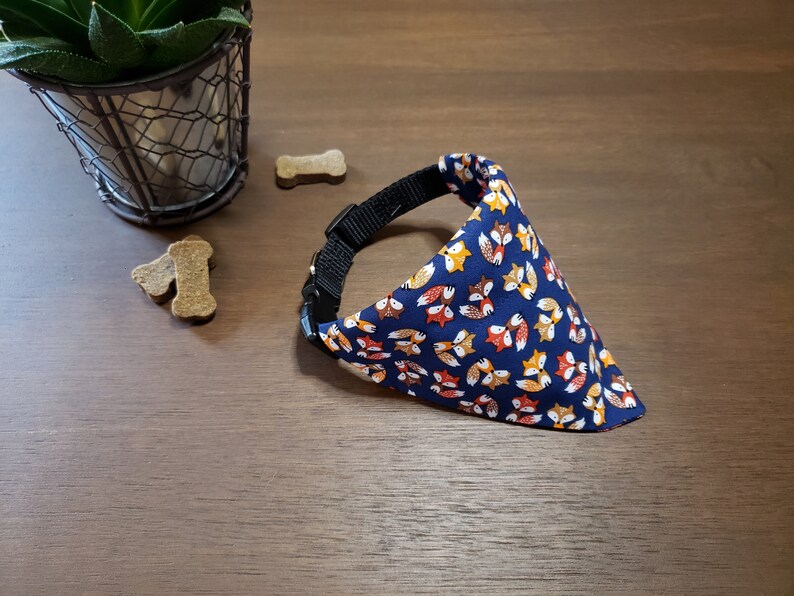 May include: A blue dog bandana with a pattern of orange, yellow, and brown foxes. The bandana is attached to a black collar.