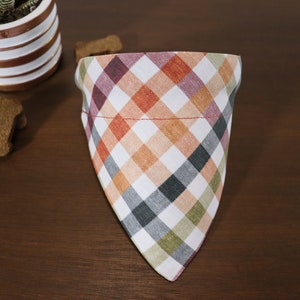 May include: A white, red, orange, green, and black plaid dog bandana. The bandana is triangular in shape and has a red stitched seam around the top edge.