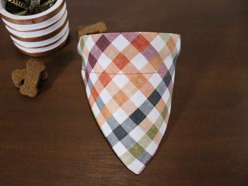 May include: A white, red, orange, green, and black plaid dog bandana. The bandana is triangular in shape and has a red stitched seam around the top edge.