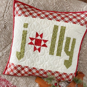 May include: A white pillow with a red and white checkered border. The pillow is quilted with a green "Jolly" design in the center. The pillow is sitting on a beige knitted blanket.