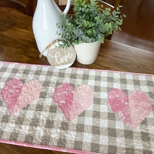 Pode incluir: Um corredor de mesa acolchoado rosa e branco com um padrão xadrez e três corações rosa. O corredor está sobre uma mesa de madeira com um vaso branco e uma planta em vaso.