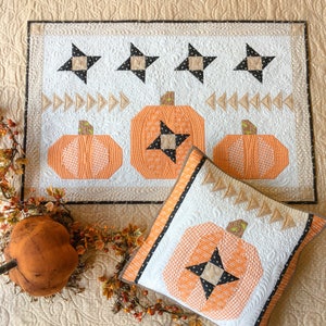 Pode incluir: Uma almofada de lançamento acolchoada branca com um design de abóbora preta e laranja. A almofada é cercada por uma borda preta e branca. A almofada está sentada em um tecido acolchoado bege com um design de abóbora.