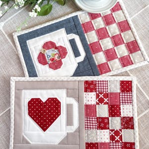 May include: Two handmade quilted mug rugs. One features a red floral design, the other a red heart. Both have a mug shape and a checkerboard pattern. The rugs are bordered with white and gray fabric.