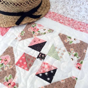 May include: A white quilt with a patchwork design featuring brown, pink, and green floral fabric. The quilt is quilted with a wavy pattern. A straw hat with a black ribbon is lying on top of the quilt.