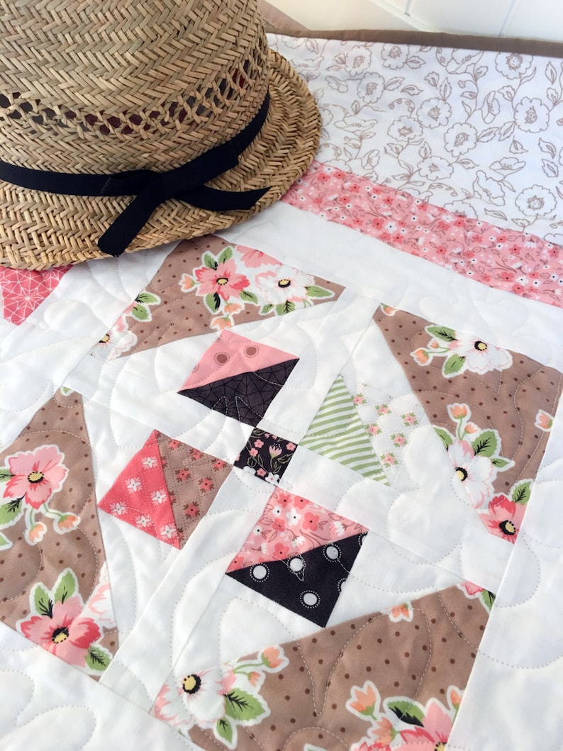 May include: A white quilt with a patchwork design featuring brown, pink, and green floral fabric. The quilt is quilted with a wavy pattern. A straw hat with a black ribbon is lying on top of the quilt.