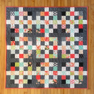 Pode incluir: Um edredom artesanal com um padrão xadrez. O edredom apresenta quadrados em várias cores, incluindo vermelho, laranja, preto, branco e estampas florais, contra um fundo cinza. Possui uma borda de bolinhas vermelhas e é exibido em um piso de madeira.