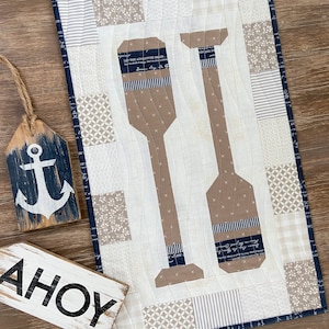 Peut inclure: Une suspension murale matelassée blanche sur le thème nautique. Le motif représente deux rames brunes avec des accents bleu et blanc rayés. Le texte "Let the adventure begin" est visible sur l'une des rames. La suspension murale est entourée d'une bordure bleu marine.