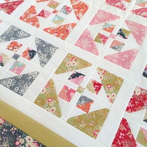 May include: A white quilt with a patchwork design featuring various floral patterns in shades of pink, red, green, and blue. The quilt is made up of squares and triangles, and the edges are trimmed with a gold ribbon.