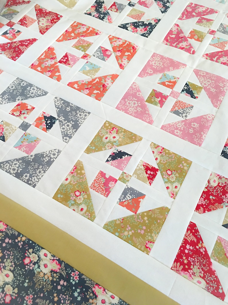 May include: A white quilt with a patchwork design featuring various floral patterns in shades of pink, red, green, and blue. The quilt is made up of squares and triangles, and the edges are trimmed with a gold ribbon.