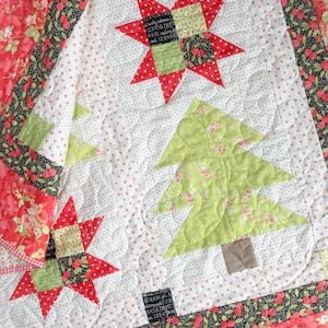 May include: A white quilt with a red, green, and black floral pattern. The quilt features a large red star in the upper right corner and two green Christmas trees in the lower left and center. The quilt is quilted with a wavy pattern.
