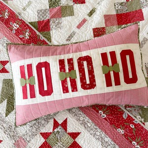 May include: A festive Christmas pillow with the words "HO HO HO" in red block letters, accented with green bows. The pillow is set on a patchwork quilt with red, green, and white patterns, creating a cozy holiday theme.