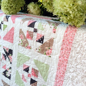 May include: A white quilt with a patchwork design featuring pink, green, and brown fabric. The quilt is quilted with a swirling pattern and has a pink and white floral border.