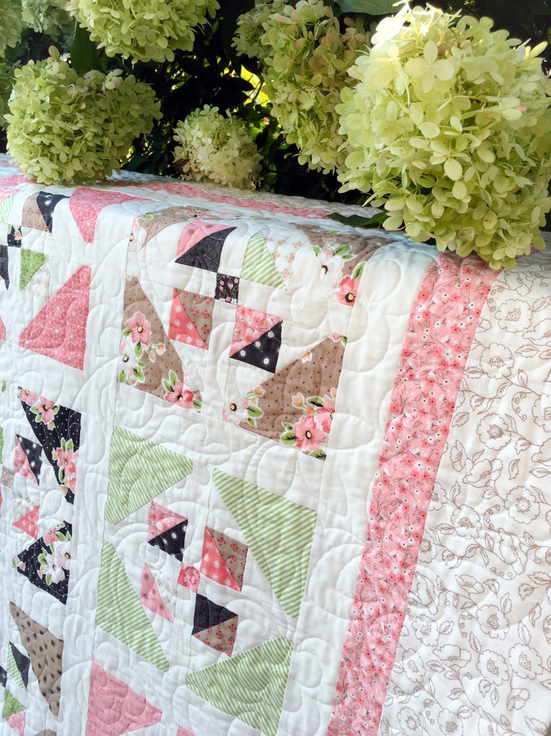 May include: A white quilt with a patchwork design featuring pink, green, and brown fabric. The quilt is quilted with a swirling pattern and has a pink and white floral border.
