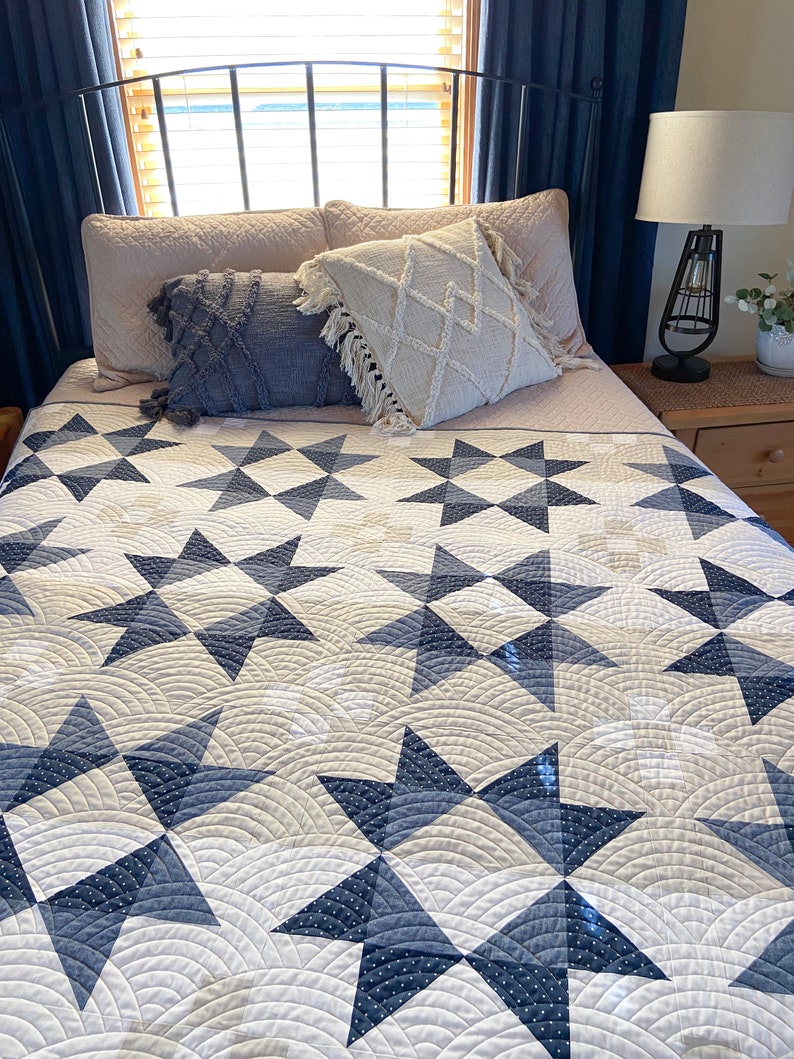May include: A white and blue quilt with a star pattern. The quilt is on a bed with a white headboard and blue curtains.