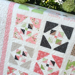 May include: A white quilt with a patchwork design featuring pink, green, and black fabric. The quilt is quilted with a swirling pattern. The quilt is displayed on a table with green foliage in the background.