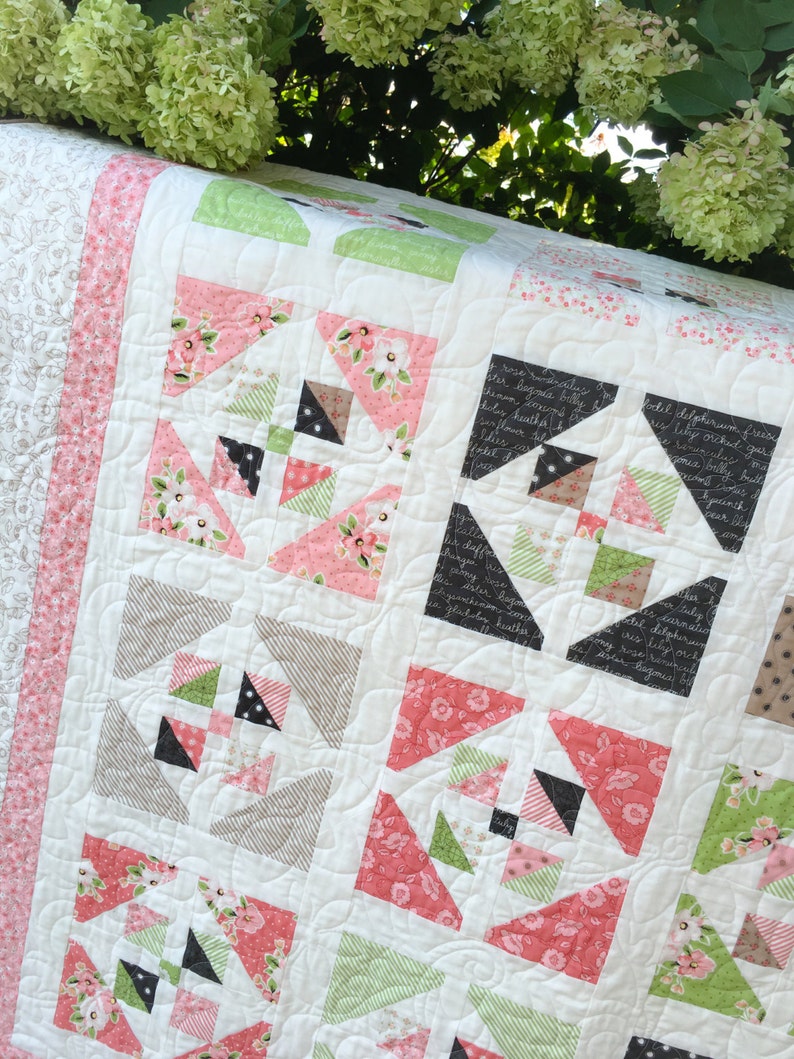 May include: A white quilt with a patchwork design featuring pink, green, and black fabric. The quilt is quilted with a swirling pattern. The quilt is displayed on a table with green foliage in the background.