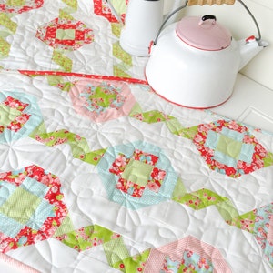 Könnte beinhalten: Ein weißer gesteppter Tischläufer mit einem floralen Muster in Rosa-, Grün- und Rottönen. Der Läufer ist mit einem gewellten Rand verziert und weist ein sich wiederholendes Muster aus Quadraten und Rauten auf. Zwei weiße Emailleteekannen mit rosafarbenen Deckeln stehen auf dem Läufer.