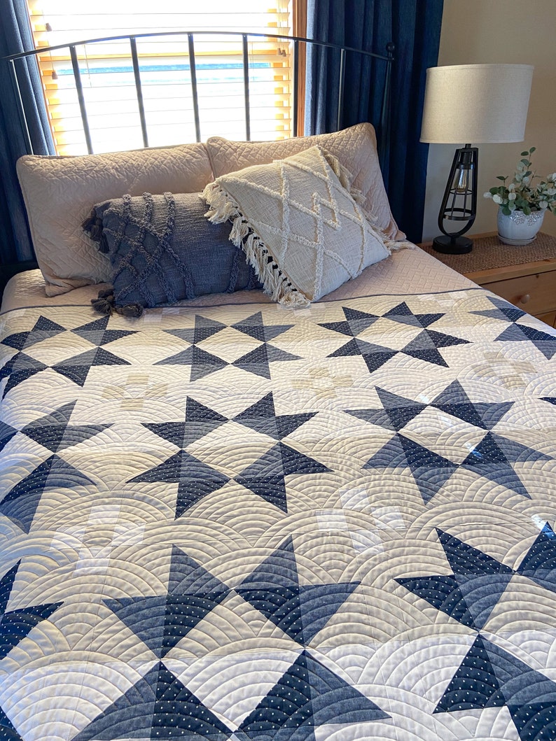 May include: A bed with a white and blue star patterned quilt. The bed has a white headboard and two pillows with fringe. A lamp is on a nightstand next to the bed.