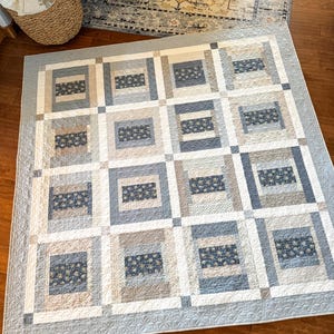May include: A square patchwork quilt featuring a repeating block pattern in shades of navy blue, light blue, beige, and white. The design consists of rectangular fabric segments with floral prints and solid textures, finished with a light blue border.