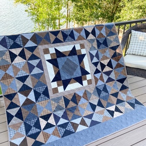 Könnte beinhalten: Eine blaue und braune Patchwork-Decke mit einem Sternmuster in der Mitte. Die Decke besteht aus verschiedenen Blautönen und braunen Stoffen, und der Stern ist aus einem dunkleren blauen Stoff gefertigt. Die Decke wird auf einem Holzdeck ausgestellt.